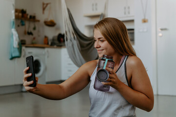 Smiling young female in sportswear holding water bottle and taking selfie on modern mobile phone during home workout