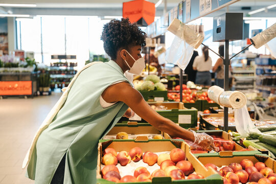Side view of young African American female in protective mask and gloves choosing red apples while visiting grocery store during coronavirus pandemic