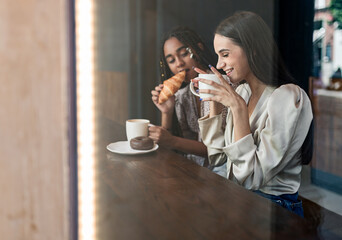 Through window view of cheerful millennial diverse girlfriends chatting happily while sitting at counter in cafe and enjoying hot coffee with pastry