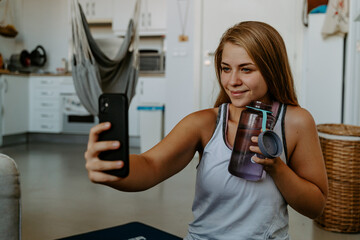 Smiling young female in sportswear holding water bottle and taking selfie on modern mobile phone during home workout