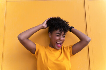 Happy young African American female with curly hair wearing bright yellow shirt and jeans standing against yellow wall and looking away