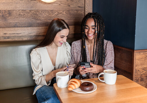 Positive young diverse female friends in stylish clothes ton smartphone while sitting at table with aromatic coffee and pastry in cozy cafe