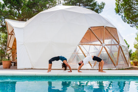 Slim partners in sportswear standing in bridge na pose while practicing yoga near swimming pool