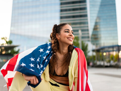Confident young ethnic lady in sports bra waving American flag and looking away against modern glass skyscraper