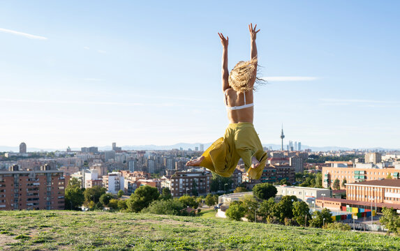 Back view full body barefoot female in bra and loose trousers jumping with arms raised while spending on grassy hilltop against city buildings