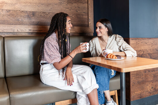 Cheerful young multiracial women in trendy outfits drinking coffee and chatting happily while spending time together in modern cafeteria