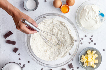 Top view crop faceless cook with whisk in hand above bowl with flour placed on table amidst baking ingredients in kitchen