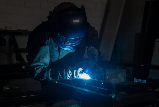 Unrecognizable male employee in protective gloves and helmet using welding machine while working in dark workshop