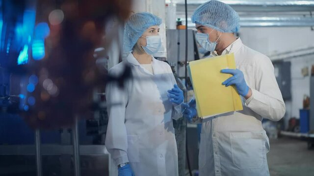Colleagues in white uniform and protective masks with papers control automatic conveyor belt in production plant workshop