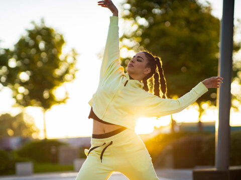 Trendy young ethnic female dancer with stylish hairstyle in activewear performing freestyle movements in park at sunset