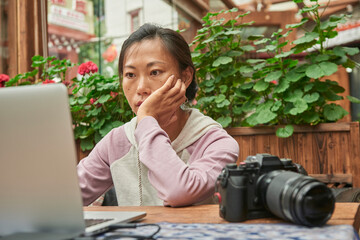 Concentrated Asian female journalist sitting at table with digital photo camera and using laptop while preparing new article