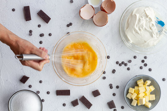 Top view crop unrecognizable cook beating eggs in bowl placed on table with baking ingredients including butter sour cream and chocolate