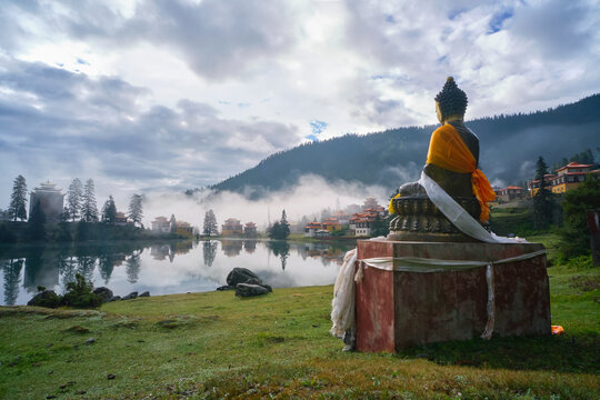 Statue of Buddha placed near peaceful Cuoka Lake located in green valley with high mountains and Tibetan Buddhist temple