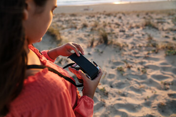 From above of crop anonymous female traveler setting vintage film camera while standing on sandy beach at sunset