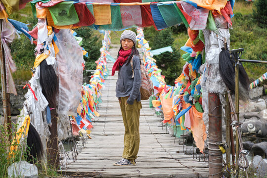 Young ethnic female tourist in warm clothing and hat looking at camera while crossing simple long narrow suspension wooden bridge decorated with bright multi colored pieces of fabric in daylight