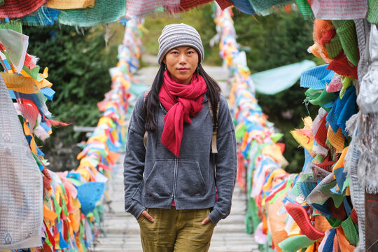 Young ethnic female tourist in warm clothing and hat looking at camera while crossing simple long narrow suspension wooden bridge decorated with bright multi colored pieces of fabric in daylight