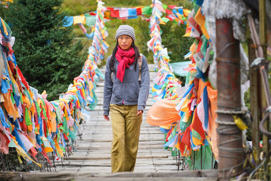 Young ethnic female tourist in warm clothing and hat looking at camera while crossing simple long narrow suspension wooden bridge decorated with bright multi colored pieces of fabric in daylight