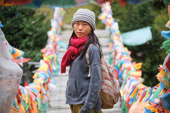 Young ethnic female tourist in warm clothing and hat looking at camera while crossing simple long narrow suspension wooden bridge decorated with bright multi colored pieces of fabric in daylight