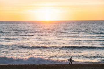Side view of anonymous surfer with surfboard walking along sandy beach near powerful waving ocean against picturesque sunset sky