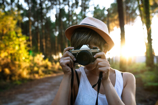 Unrecognizable female photographer in casual outfit and hat standing on path in green forest and taking pictures on sunny day