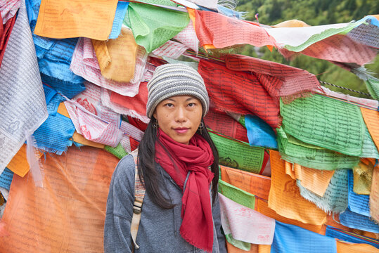 Young ethnic female tourist in warm clothing and hat looking at camera while crossing simple long narrow suspension wooden bridge decorated with bright multi colored pieces of fabric in daylight