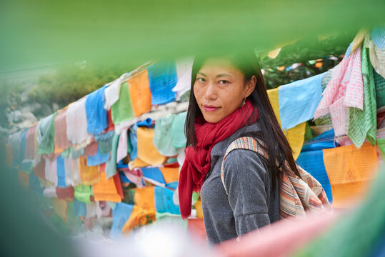 Young ethnic female tourist in warm clothing and hat looking at camera while crossing simple long narrow suspension wooden bridge decorated with bright multi colored pieces of fabric in daylight