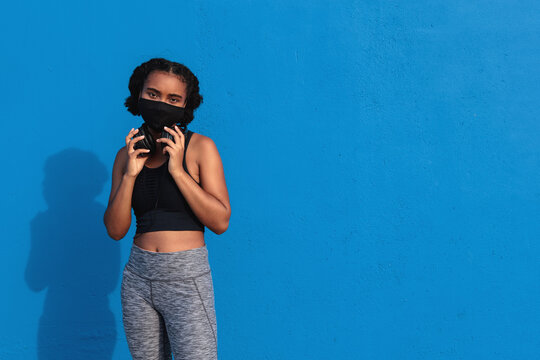 Young African American female athlete with headphones on neck in face mask and sportswear standing against blue background and looking at camera