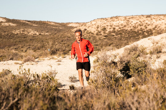 Front view of active male jogger running uphill in sandy semi desert terrain during workout in sunny morning