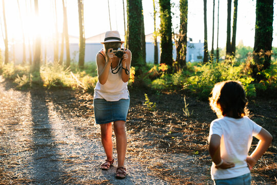 Unrecognizable young female photographer in casual clothes and hat shooting adorable little son standing on alley in picturesque park