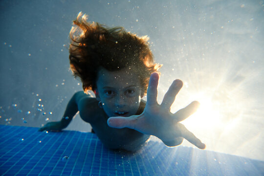 Underwater shot of cheerful teenage boy diving into blue swimming pool water with eyes closed and smiling happily