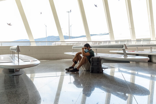 Side view of discouraged male tourist in protective mask sitting on floor in departure lounge of airport with phone while waiting for flight during coronavirus pandemic