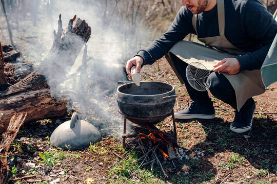Crop anonymous male cook in apron preparing lunch while putting pasty in boiling oil in pot on campfire