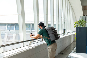 Side view of male tourist wearing medical mask standing in departure lounge of airport and browsing cellphone