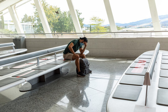 Side view of discouraged male tourist in protective mask sitting in departure lounge of airport with phone while waiting for flight during coronavirus pandemic