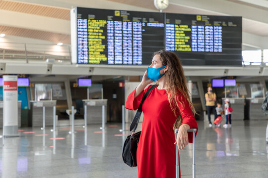 Serious female traveler in mask standing with suitcase on background of departure board and talking on the phone while waiting for flight