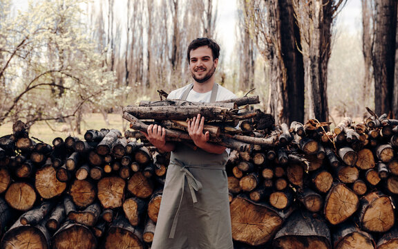 Man in apron taking logs from pile of firewood in forest for picnic