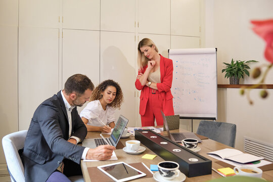 Cheerful coworkers gathering in meeting room and sharing ideas about marketing strategy while working with laptop and taking notes on flipchart