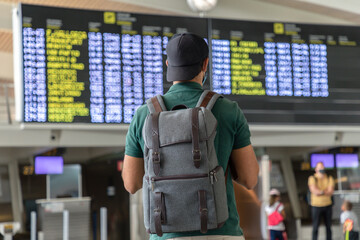 Back view of unrecognizable male traveler with backpack checking schedule on departure board while standing in international airport and waiting for flight