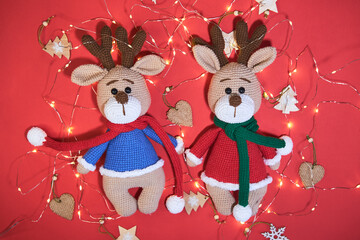 Flat background on Valentines day or new year. Two adorable hand knitted amigurumi reindeer in red and blue sweater and scarves on red background with bright yellow garland and Christmas tree toys.