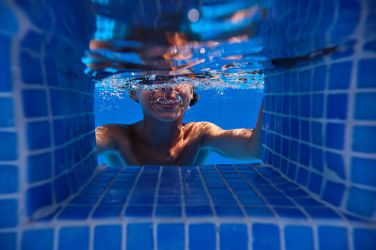 Underwater shot of cheerful teenage boy diving into blue swimming pool water with eyes closed and smiling happily