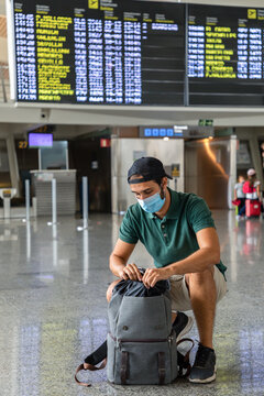 Calm male tourist squatting against departure board in airport and zipping his backpack while waiting for flight