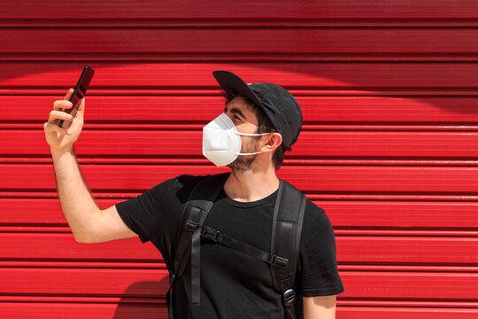 Young male in respiratory mask taking selfie on cellphone near red wall standing on the street during COVID 19 pandemic
