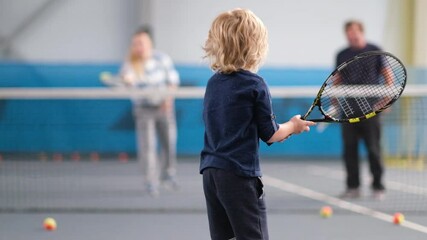 A female coach teaches a little boy to play on an indoor court. A professional tennis instructor tosses the ball to hit the racket to a child, how to hit the ball with a racket