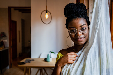 Pensive young African American woman in trendy eyeglasses covering half of her body with curtain and looking at camera at home