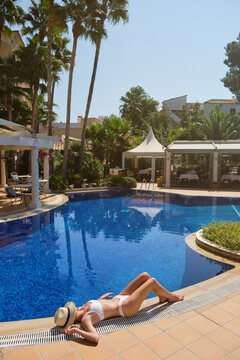 High angle of unrecognizable young slim lady in swimwear and straw hat sunbathing while lying near swimming pool during summer vacation