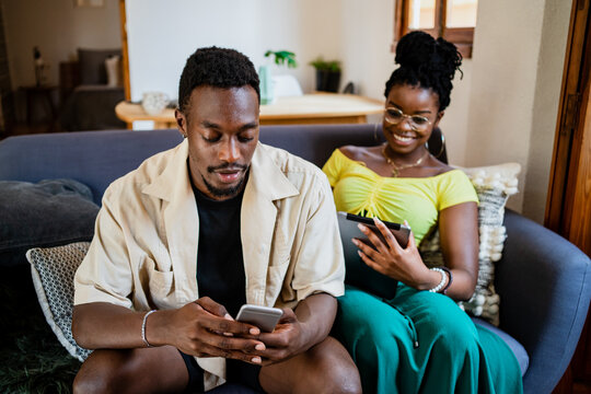Content young African American lady reading information on tablet while resting on sofa with boyfriend using smartphone at home