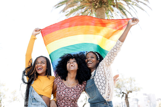 Low angle of delighted African American females in love hugging on street while standing with LGBT rainbow flag and looking away