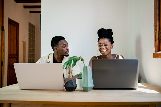 Front view of positive young African American man and woman in casual clothes and smiling while working together on laptops at home - Powered by Adobe