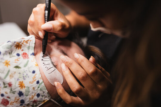 From above of crop anonymous female master using eyeliner and patch while preparing client for eyelash extension in beauty salon
