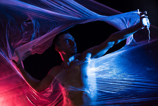 Aggressive male in casual clothing shouting and looking at camera while ripping transparent tape in light of red and blue lamps